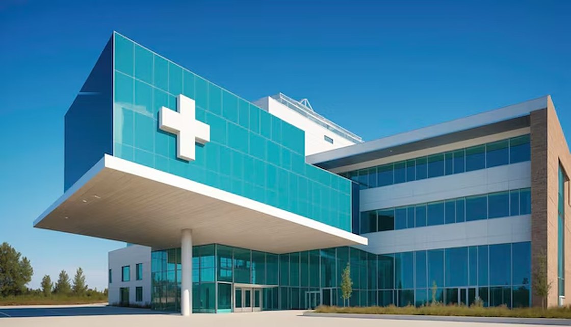 Extérieur d’une clinique moderne en plein jour : une façade de verre turquoise portant une croix blanche au-dessus d’un auvent d’entrée en porte-à-faux.